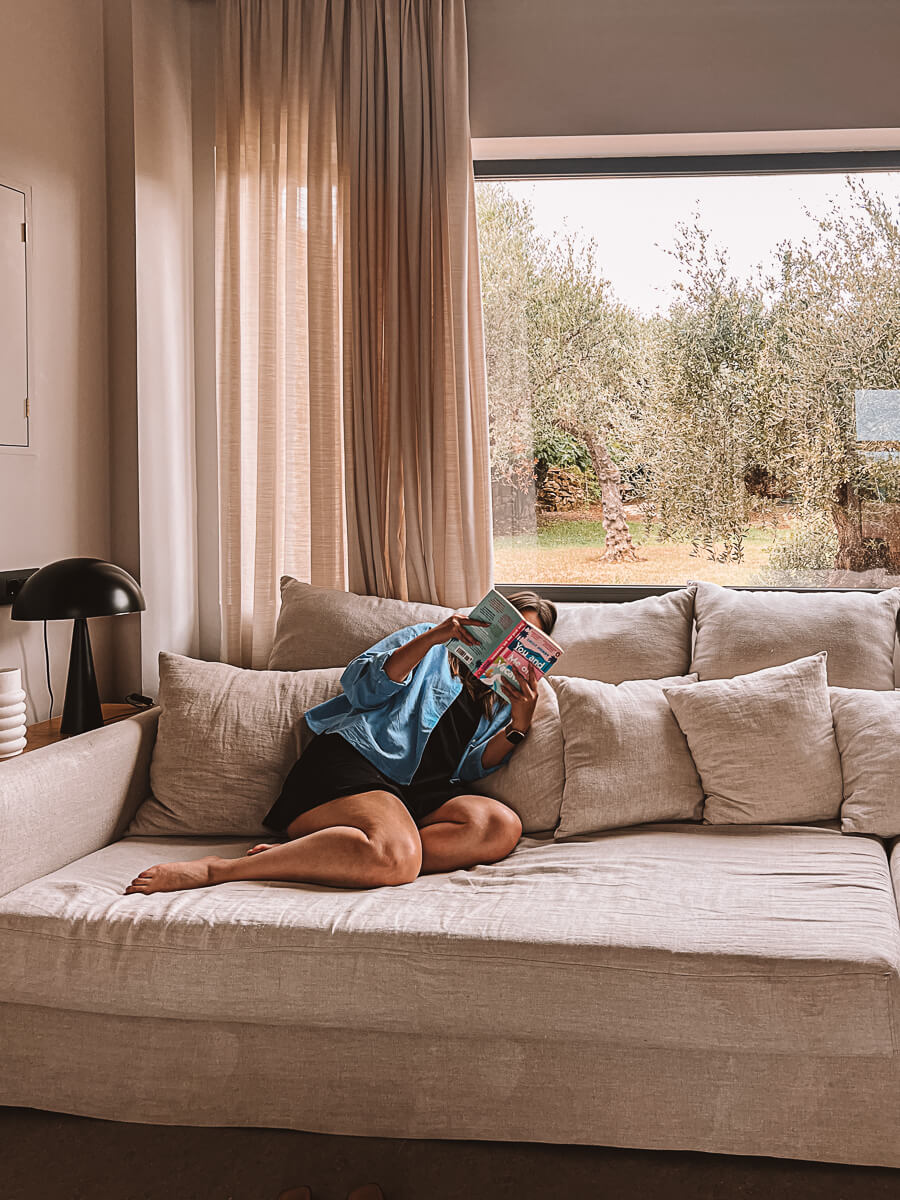 Girl reading on cosy sofa indoors on reading retreat in Greece with Aweventurer. There are olive trees outside the window. 
