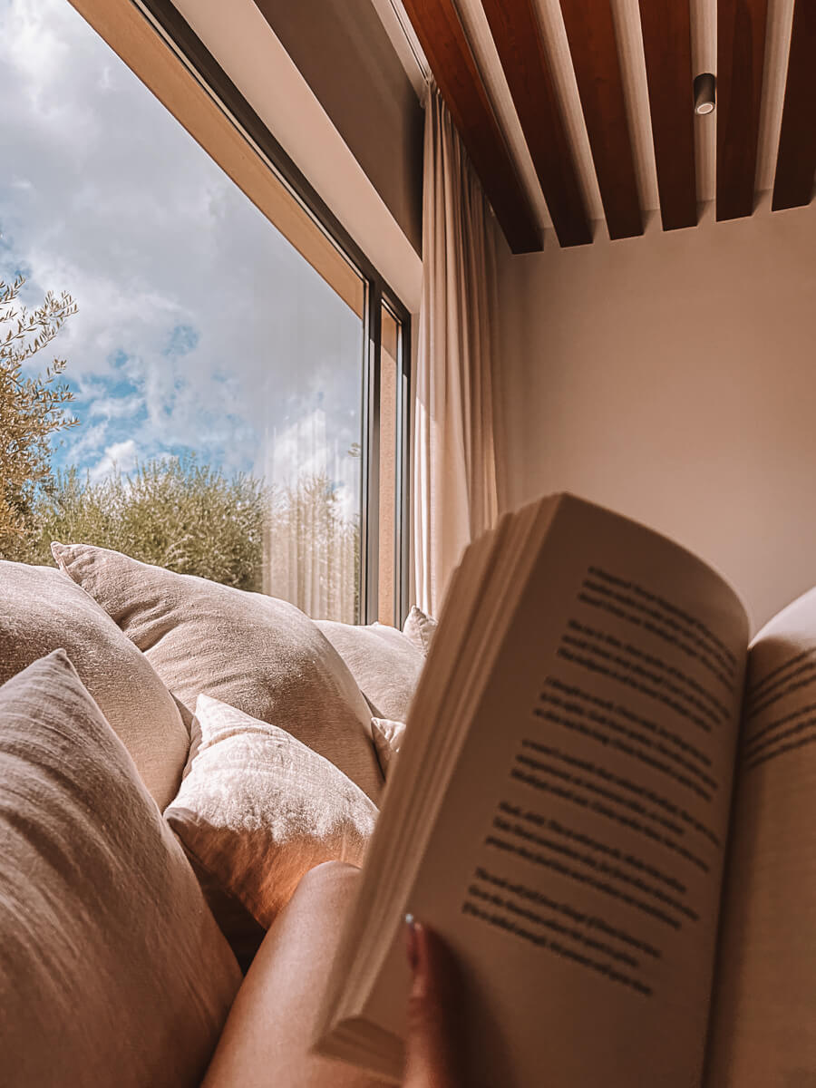 Girl reading on cosy sofa in Cielo Villas on reading retreatin Zakynthos Greece with view of olive trees outside of the window. 